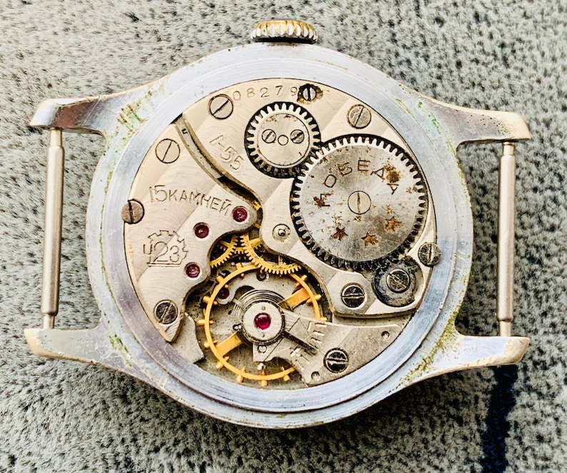 Puede incluir: Primer plano del mecanismo de un reloj vintage. La caja plateada enmarca los intrincados engranajes, joyas y muelles. La esfera del reloj muestra el texto "15 KAMHEЙ" y "423". El reloj tiene una peque&ntilde;a corona.