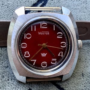 Könnte beinhalten: Eine silberne Armbanduhr mit rotem Zifferblatt und weißen Ziffern. Die Uhr hat ein quadratisches Gehäuse und ein Metallband. Das Zifferblatt der Uhr zeigt "Wostok" in weißen Buchstaben.