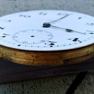 Puede incluir: Primer plano de una esfera de reloj de bolsillo vintage. La esfera blanca presenta n&uacute;meros negros, un peque&ntilde;o dial de segundos y agujas negras. La caja de metal dorado del reloj es visible, mostrando signos de edad.