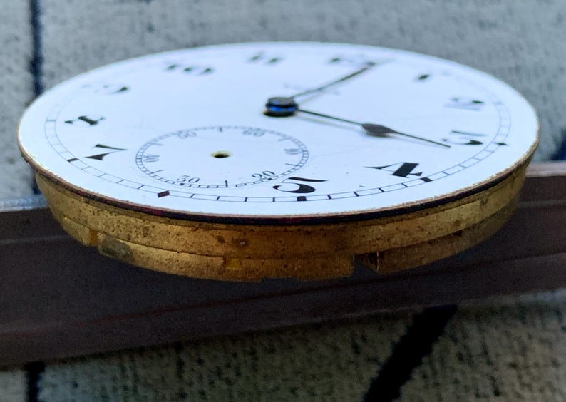 Puede incluir: Primer plano de una esfera de reloj de bolsillo vintage. La esfera blanca presenta n&uacute;meros negros, un peque&ntilde;o dial de segundos y agujas negras. La caja de metal dorado del reloj es visible, mostrando signos de edad.
