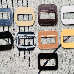 May include: A collection of eight rectangular belt buckles in various colors, including blue, beige, brown, silver, black, and yellow. Each buckle has a rectangular opening with three horizontal bars.