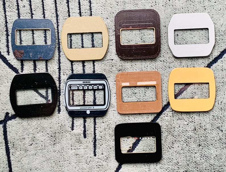 May include: A collection of eight rectangular belt buckles in various colors, including blue, beige, brown, silver, black, and yellow. Each buckle has a rectangular opening with three horizontal bars.