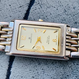 May include: A rectangular Perucci watch with a gold-tone face and hands, and gold-tone hour markers. The watch has a silver-tone case and a two-tone silver and gold band. The watch face includes the text "Perucci" and "Expression Water Proof".