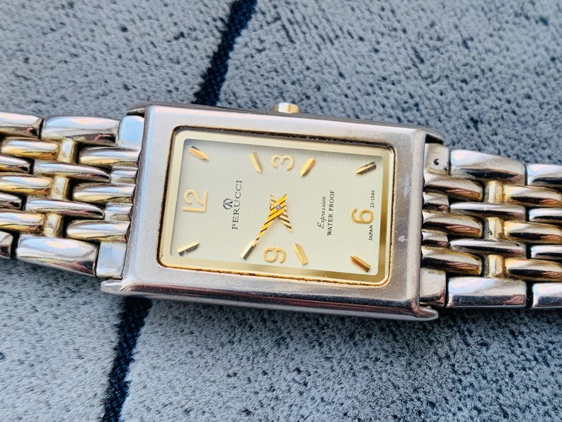 May include: A rectangular Perucci watch with a gold-tone face and hands, and gold-tone hour markers. The watch has a silver-tone case and a two-tone silver and gold band. The watch face includes the text "Perucci" and "Expression Water Proof".