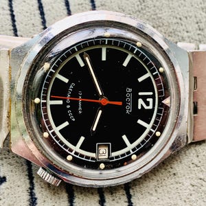 Puede incluir: Un reloj de pulsera plateado con una esfera negra y una correa de metal plateado. La esfera del reloj tiene marcas blancas y una aguja de segundos roja. La esfera del reloj también tiene el texto "Восток" y "17 камней" en cirílico.