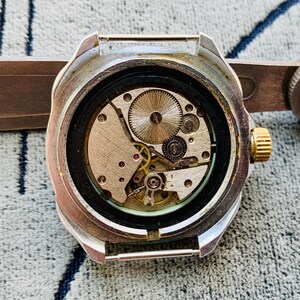 May include: Close-up of a vintage wristwatch with the back removed, revealing the intricate mechanical movement. The watch has a silver-colored case, a gold-colored crown, and a black bezel. The internal gears and components are visible.
