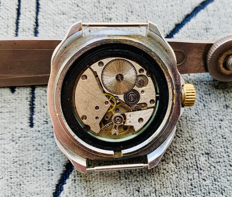 May include: Close-up of a vintage wristwatch with the back removed, revealing the intricate mechanical movement. The watch has a silver-colored case, a gold-colored crown, and a black bezel. The internal gears and components are visible.