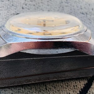Puede incluir: Primer plano de un reloj de pulsera vintage con caja plateada y cristal abombado transparente. La esfera del reloj es de color oro claro. El reloj descansa sobre una superficie oscura y texturizada.