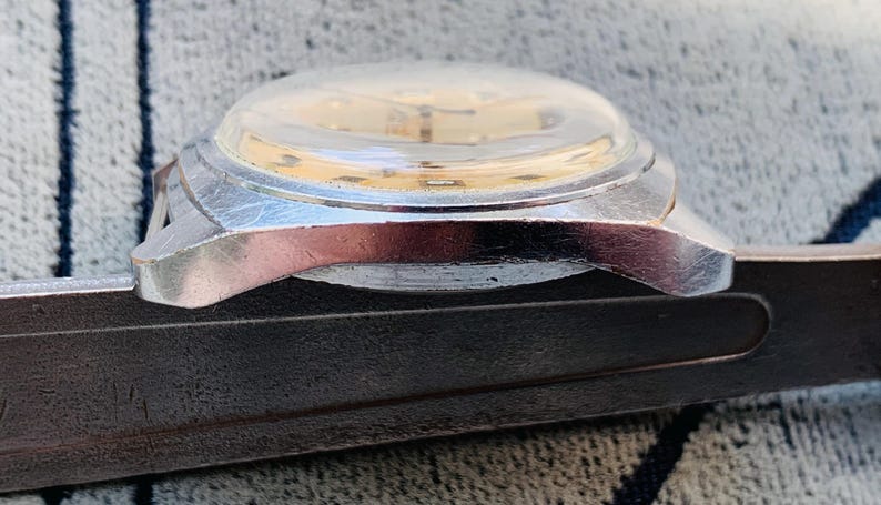 Puede incluir: Primer plano de un reloj de pulsera vintage con caja plateada y cristal abombado transparente. La esfera del reloj es de color oro claro. El reloj descansa sobre una superficie oscura y texturizada.