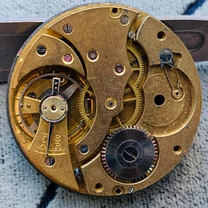 Puede incluir: Primer plano de un movimiento de reloj antiguo, que muestra intrincados engranajes de lat&oacute;n, tornillos y componentes. El mecanismo circular presenta una compleja disposici&oacute;n de engranajes entrelazados, peque&ntilde;as joyas de rub&iacute; y un engranaje plateado. La paleta de colores es dorada y plateada.