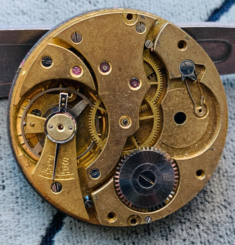 Puede incluir: Primer plano de un movimiento de reloj antiguo, que muestra intrincados engranajes de lat&oacute;n, tornillos y componentes. El mecanismo circular presenta una compleja disposici&oacute;n de engranajes entrelazados, peque&ntilde;as joyas de rub&iacute; y un engranaje plateado. La paleta de colores es dorada y plateada.