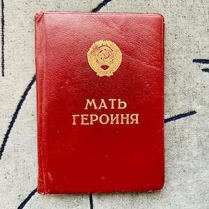 May include: A red book with gold lettering and a gold emblem. The text on the cover reads "MATЬ ГЕРОИНЯ". The book is rectangular and appears to be made of leather. The emblem is a gold star with a hammer and sickle.