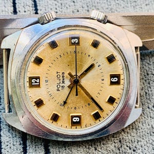 Puede incluir: Primer plano de un reloj de pulsera vintage Poljot 18 jewels. El reloj tiene una esfera dorada con manecillas y marcadores negros. La caja plateada presenta dos coronas y una forma rectangular.
