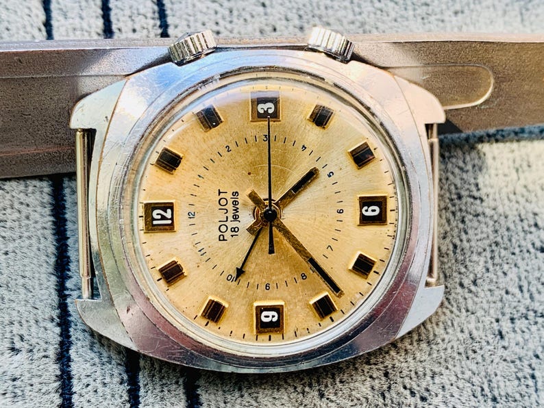 Puede incluir: Primer plano de un reloj de pulsera vintage Poljot 18 jewels. El reloj tiene una esfera dorada con manecillas y marcadores negros. La caja plateada presenta dos coronas y una forma rectangular.