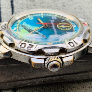 Op de afbeelding: Close-up van een zilveren polshorloge met een blauwe en groene wijzerplaat. Het horloge heeft een ronde wijzerplaat met witte cijfers en een rode secondewijzer. Het horloge heeft een metalen band.