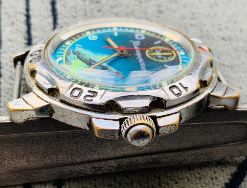 Op de afbeelding: Close-up van een zilveren polshorloge met een blauwe en groene wijzerplaat. Het horloge heeft een ronde wijzerplaat met witte cijfers en een rode secondewijzer. Het horloge heeft een metalen band.