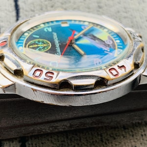Op de afbeelding: Close-up van een zilverkleurig polshorloge met een blauwe en turquoise wijzerplaat. De wijzerplaat heeft een kompasontwerp en een vliegtuig. De bezel heeft rode en gouden markeringen. De wijzers van het horloge zijn rood en goud.