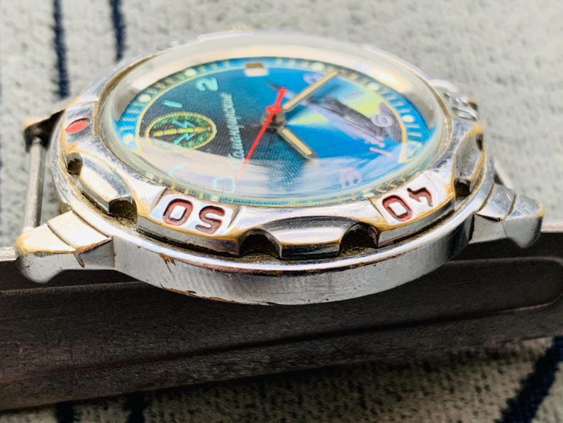 Op de afbeelding: Close-up van een zilverkleurig polshorloge met een blauwe en turquoise wijzerplaat. De wijzerplaat heeft een kompasontwerp en een vliegtuig. De bezel heeft rode en gouden markeringen. De wijzers van het horloge zijn rood en goud.
