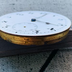 Puede incluir: Primer plano de una esfera de reloj vintage con esfera blanca, n&uacute;meros negros y delicadas manecillas. El reloj tiene un bisel dorado y una esfera secundaria m&aacute;s peque&ntilde;a. El reloj est&aacute; sobre una superficie met&aacute;lica.