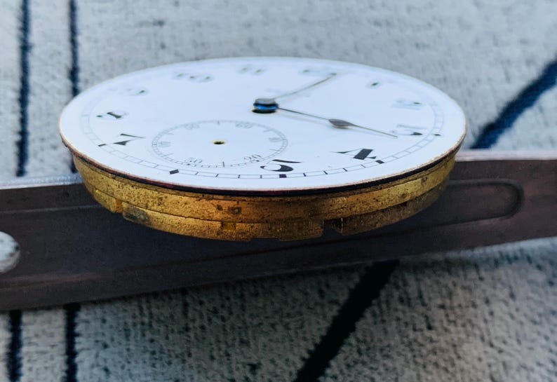 Puede incluir: Primer plano de una esfera de reloj vintage con esfera blanca, n&uacute;meros negros y delicadas manecillas. El reloj tiene un bisel dorado y una esfera secundaria m&aacute;s peque&ntilde;a. El reloj est&aacute; sobre una superficie met&aacute;lica.