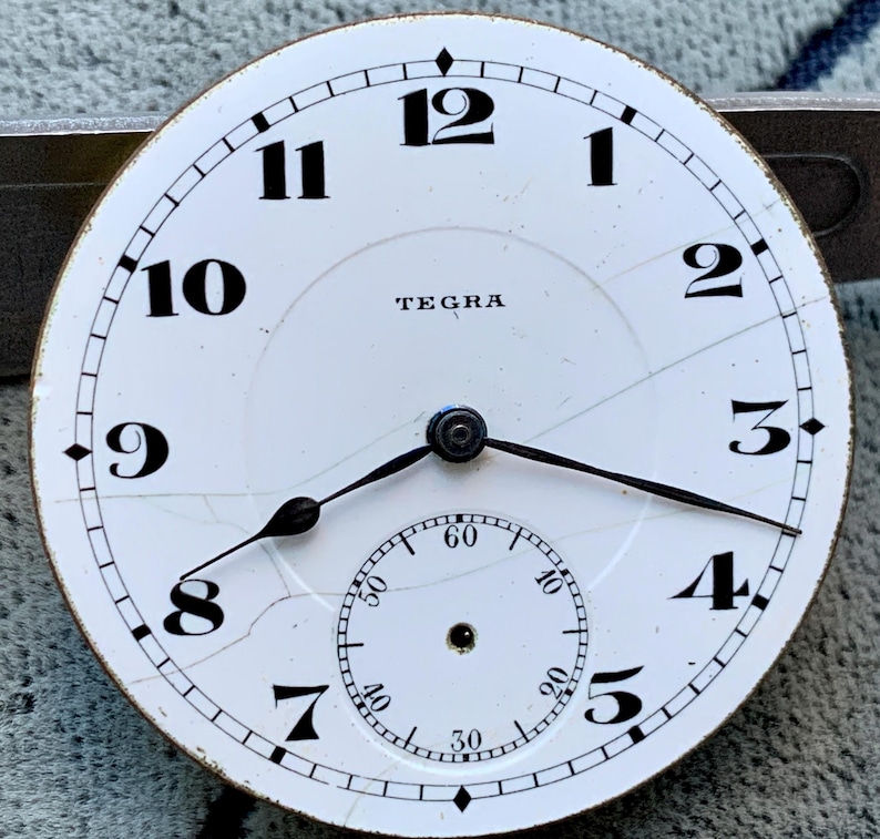 Puede incluir: Una esfera de reloj blanca con n&uacute;meros y manecillas negras. La esfera tiene una esfera secundaria m&aacute;s peque&ntilde;a en la parte inferior y la palabra "TEGRA" en la parte superior. La esfera del reloj est&aacute; agrietada.