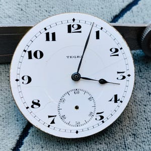 Puede incluir: Una esfera de reloj blanca con n&uacute;meros y manecillas negras, con un peque&ntilde;o dial de segundos. La palabra "TEGRA" est&aacute; impresa en la esfera. La esfera del reloj est&aacute; sobre un fondo met&aacute;lico.
