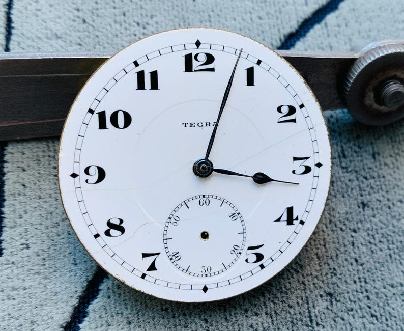 Puede incluir: Una esfera de reloj blanca con n&uacute;meros y manecillas negras, con un peque&ntilde;o dial de segundos. La palabra "TEGRA" est&aacute; impresa en la esfera. La esfera del reloj est&aacute; sobre un fondo met&aacute;lico.