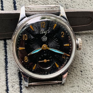 May include: A black and silver wristwatch with a black face and gold accents. The watch has a blue hand and a crown with a star on it. The watch face has the text "СССР" on it.
