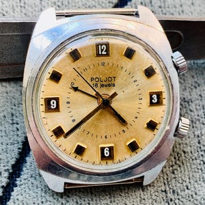 Puede incluir: Un reloj de pulsera vintage con esfera dorada y caja plateada. La esfera muestra la marca "POLJOT" y "18 jewels". Las manecillas y los marcadores de hora son negros. El reloj tiene dos botones laterales y una fijaci&oacute;n de correa met&aacute;lica.