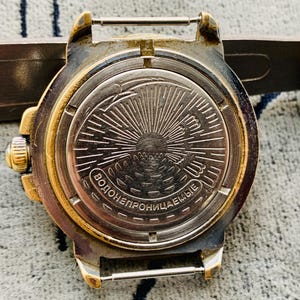 Op de afbeelding: Close-up van de achterkant van een vintage polshorloge. Het horloge heeft een goudkleurige bezel en een zilverkleurige achterkant met een sunburst-ontwerp. De tekst "ВОДОНЕПРОНИЦАЕМЫЕ" is zichtbaar op de achterkant van het horloge.