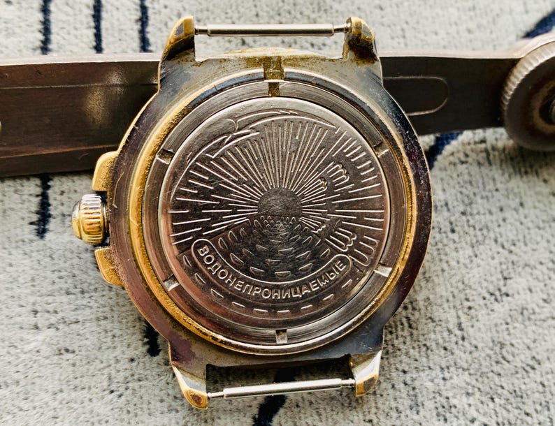 Op de afbeelding: Close-up van de achterkant van een vintage polshorloge. Het horloge heeft een goudkleurige bezel en een zilverkleurige achterkant met een sunburst-ontwerp. De tekst "ВОДОНЕПРОНИЦАЕМЫЕ" is zichtbaar op de achterkant van het horloge.