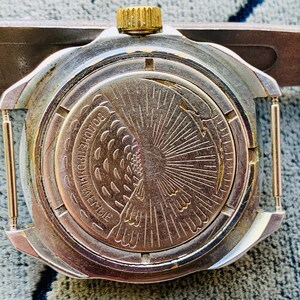 May include: Close-up of a vintage silver-toned watch back with a gold crown. The watch face features an engraved design of a bird and sunburst pattern. The watch is attached to a silver-toned metal tool.