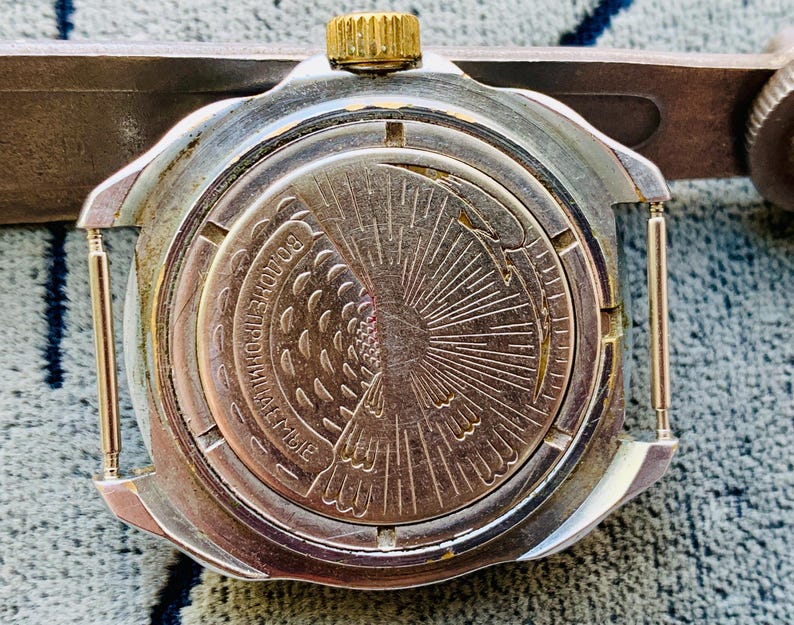 May include: Close-up of a vintage silver-toned watch back with a gold crown. The watch face features an engraved design of a bird and sunburst pattern. The watch is attached to a silver-toned metal tool.