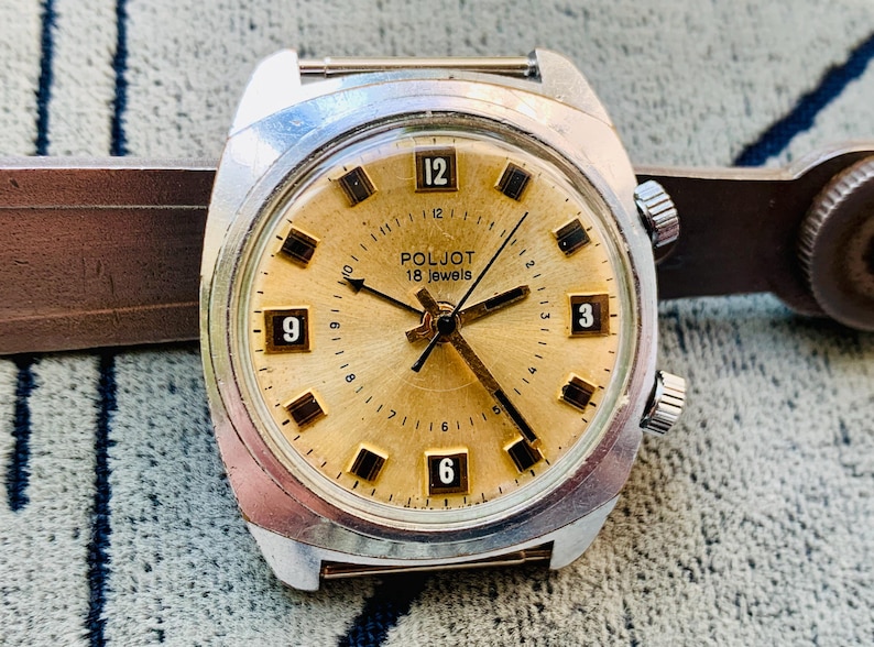 Puede incluir: Un reloj de pulsera vintage Poljot 18 jewels con esfera dorada y caja plateada. La esfera muestra marcadores de hora rectangulares negros y los n&uacute;meros 3, 6, 9 y 12. El reloj est&aacute; sujeto a una correa de cuero marr&oacute;n.