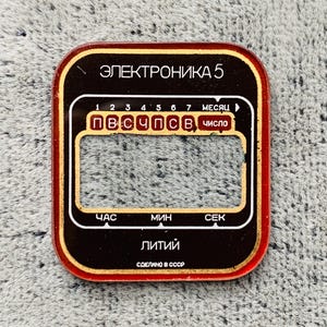 May include: A rectangular, vintage watch face with a red border and gold accents. The face features Cyrillic text, including "ЭЛЕКТРОНИКА 5" and "Литий". It has a display area and labels for hours, minutes, and seconds.