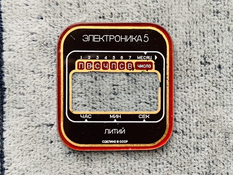 May include: A rectangular, vintage watch face with a red border and gold accents. The face features Cyrillic text, including "ЭЛЕКТРОНИКА 5" and "Литий". It has a display area and labels for hours, minutes, and seconds.