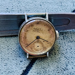 Puede incluir: Un reloj de pulsera Doxa vintage con una esfera de color crema y agujas negras. El reloj tiene una caja plateada y una correa de cuero negra. La esfera del reloj tiene las palabras "Doxa Anti-Magnetic" impresas.