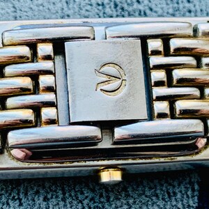 May include: Close-up of a watch band with a woven design. The band features alternating gold and silver-toned rectangular links. A silver-toned clasp with a gold emblem is centered. The watch band is a stylish accessory.
