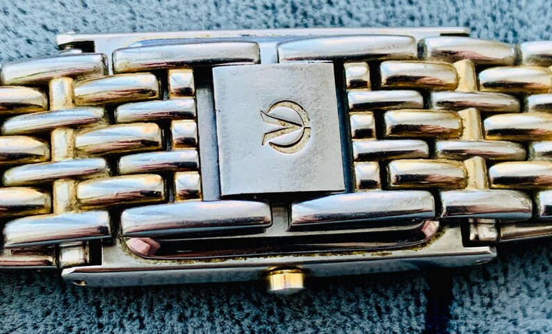 May include: Close-up of a watch band with a woven design. The band features alternating gold and silver-toned rectangular links. A silver-toned clasp with a gold emblem is centered. The watch band is a stylish accessory.