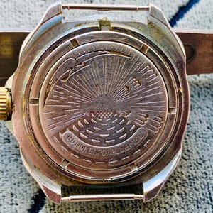 May include: Close-up of a vintage silver-toned wristwatch back. The watch features a detailed engraved design of a sunburst with a bird and text. The watch has a gold-colored crown and is attached to a brown leather strap.