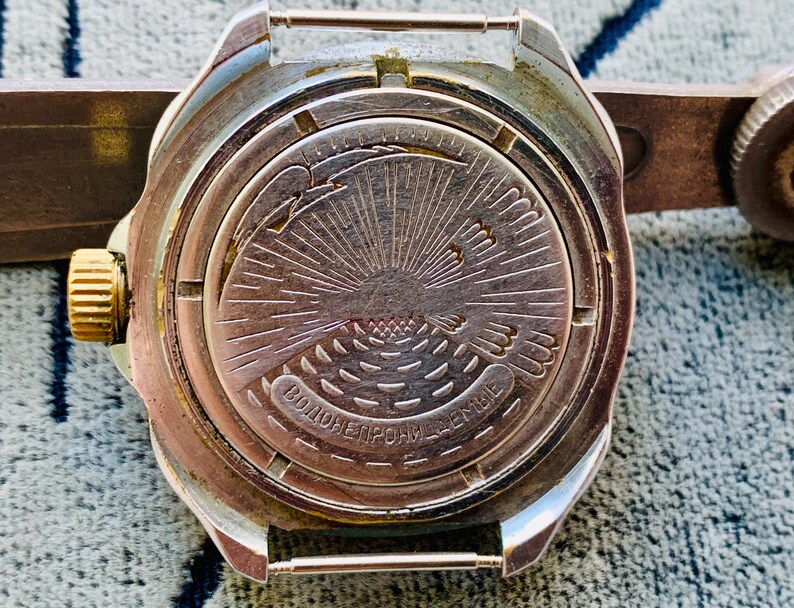 May include: Close-up of a vintage silver-toned wristwatch back. The watch features a detailed engraved design of a sunburst with a bird and text. The watch has a gold-colored crown and is attached to a brown leather strap.