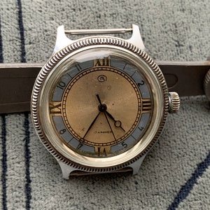 Könnte beinhalten: Eine silberne Armbanduhr mit einem runden Zifferblatt und schwarzen Zeigern. Die Uhr hat römische Ziffern und einen kleinen Sekundenzeiger. Das Zifferblatt der Uhr ist hellbraun mit einem blauen Außenring. Die Uhr ist an einem braunen Lederarmband.