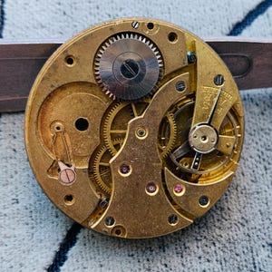 Puede incluir: Primer plano de un movimiento de reloj antiguo con intrincados engranajes, ruedas y joyas. Los componentes de color lat&oacute;n est&aacute;n dispuestos en un patr&oacute;n circular, con un engranaje plateado en el centro. El mecanismo del reloj est&aacute; sujeto por una herramienta plateada.