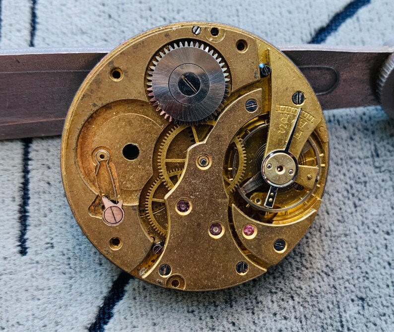 Puede incluir: Primer plano de un movimiento de reloj antiguo con intrincados engranajes, ruedas y joyas. Los componentes de color lat&oacute;n est&aacute;n dispuestos en un patr&oacute;n circular, con un engranaje plateado en el centro. El mecanismo del reloj est&aacute; sujeto por una herramienta plateada.