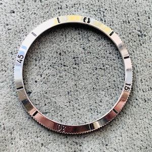 Pode incluir: Um aro de relógio prateado com marcas pretas. O aro tem números de 0 a 60, com marcas a cada 5 minutos. O aro é projetado para um relógio com um diâmetro de aproximadamente 40 mm.