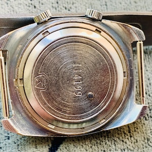 Puede incluir: Primer plano de la parte trasera de un reloj de pulsera vintage. El reloj tiene una caja de metal plateado con dos peque&ntilde;as coronas texturizadas. La parte trasera del reloj est&aacute; grabada con el n&uacute;mero "114199" y un s&iacute;mbolo. El reloj est&aacute; sujeto a una correa marr&oacute;n.