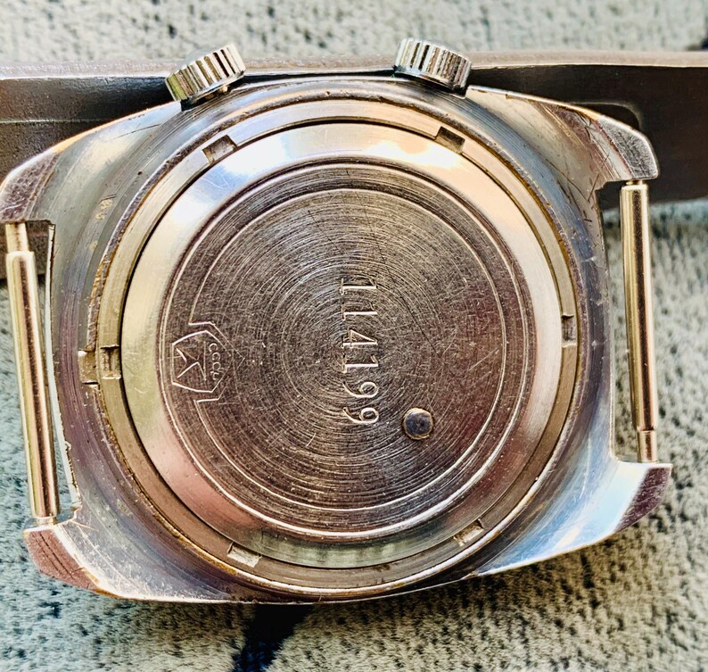 Puede incluir: Primer plano de la parte trasera de un reloj de pulsera vintage. El reloj tiene una caja de metal plateado con dos peque&ntilde;as coronas texturizadas. La parte trasera del reloj est&aacute; grabada con el n&uacute;mero "114199" y un s&iacute;mbolo. El reloj est&aacute; sujeto a una correa marr&oacute;n.