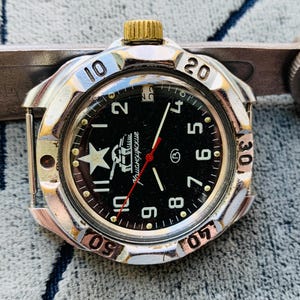 May include: A vintage wristwatch with a black face, white star, and gold-colored crown. The watch has a silver-colored bezel with black numerals and a red second hand. The watch is approximately 1.5 inches in diameter.