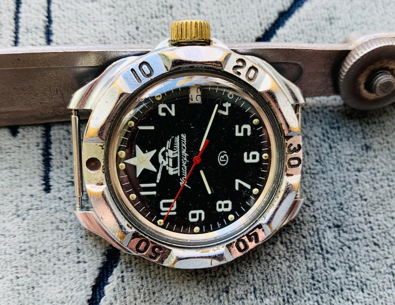 May include: A vintage wristwatch with a black face, white star, and gold-colored crown. The watch has a silver-colored bezel with black numerals and a red second hand. The watch is approximately 1.5 inches in diameter.
