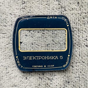 May include: A dark blue, rectangular watch face cover with a gold-colored rectangular window in the center. The text on the cover is in Cyrillic script, with the words "DATA - SEC", "ELECTROHIKA 5", and "MADE IN USSR".